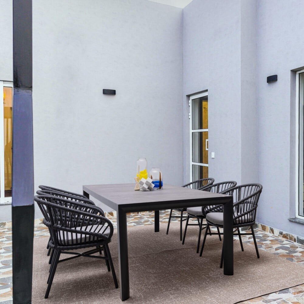 Alt: Spazio esterno con tavolo e sedie in rattan nero su pavimento in ciottoli.