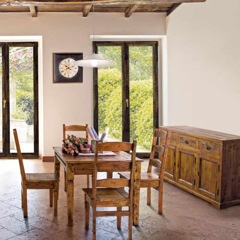 Ampio soggiorno rustico con tavolo in legno, sedie e grandi finestre che affacciano sul verde outdoor.