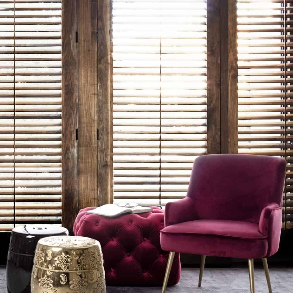 Sedia in velluto rosa con piantana decorativa, vicino a tappeto e tendaggi in legno, atmosfera elegante.