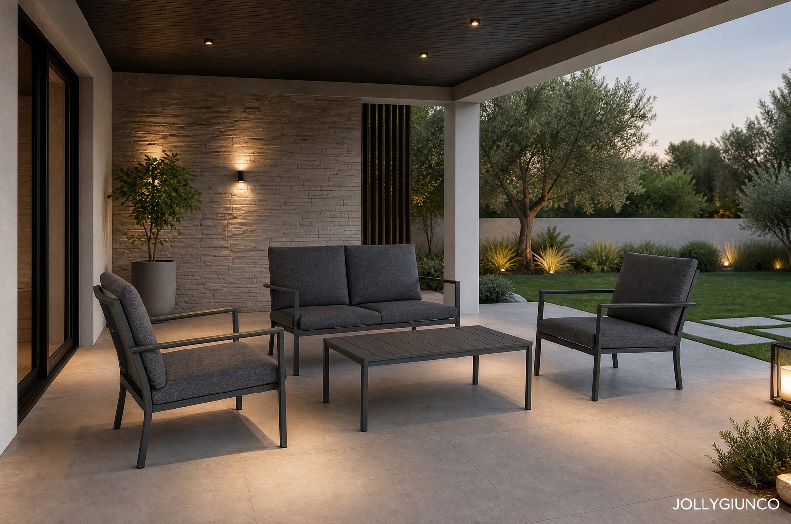 SET4 SALOTTO C-C KONNOR ANTRACITE CX2310 - JOLLYGIUNCO Modern outdoor patio with gray cushioned seating, a low coffee table, stone wall, and a landscaped garden with trees at dusk.