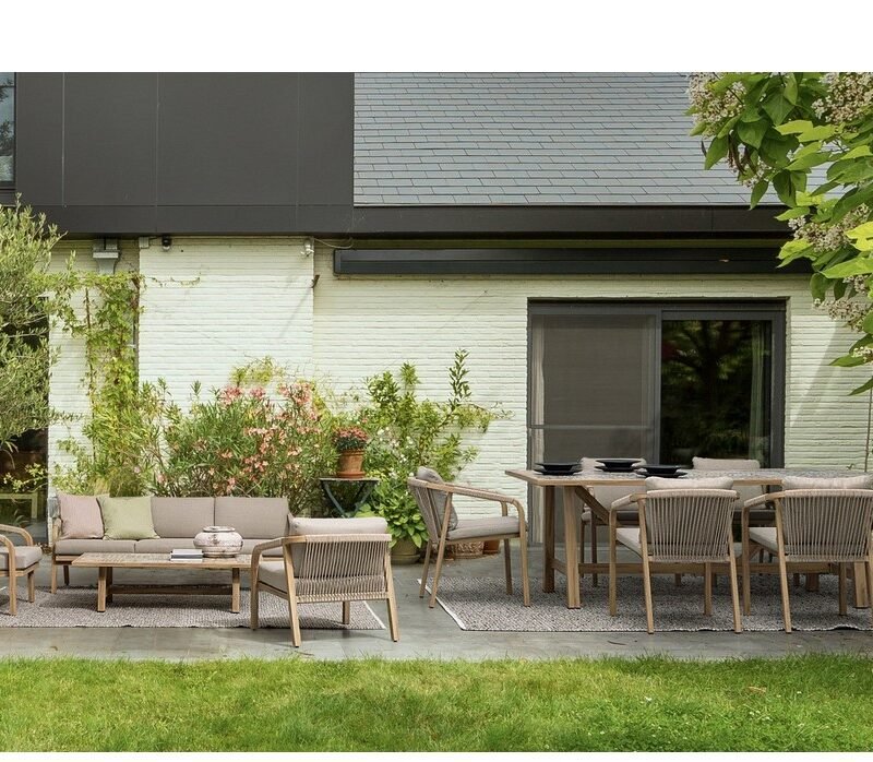 Outdoor patio with a wooden dining table and chairs on a tiled deck, surrounded by green plants and a white brick wall in the background.