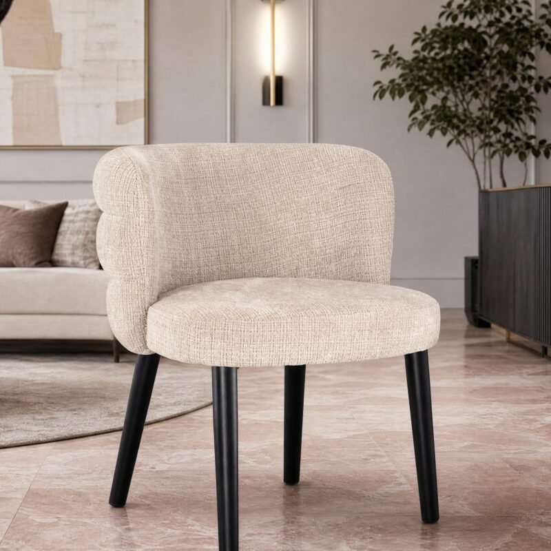 Beige upholstered chair with black legs in a modern living room, with a cream sofa, abstract wall art, and a gold lamp on a dark cabinet in the background.