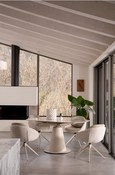 TAV ALJAMI 160X102-160 CREMA-STATUARIO7 - JOLLYGIUNCO Bright modern dining area with a round glass table, six cream cushioned chairs, a fireplace, and large windows overlooking trees.