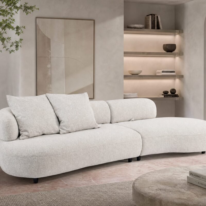 Neutral-toned living room with a plush beige sectional, potted tree on the left, and built-in lit shelves on the right.