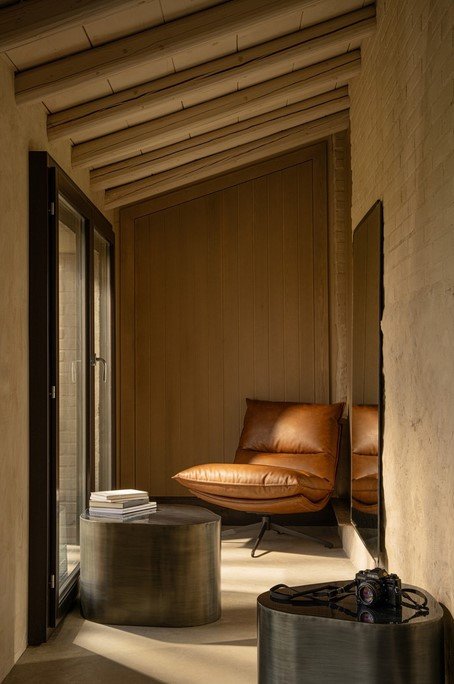 POLTRONA ALANEZ COGNAC 4 - JOLLYGIUNCO Cozy reading nook with a caramel leather lounge chair, stack of books, and round metal tables in a sunlit hallway.