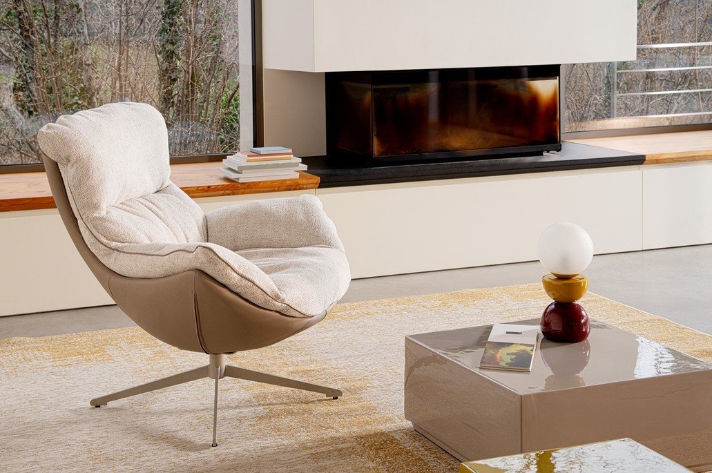 POLTRONA ARMELA BEIGE 11 - JOLLYGIUNCO Cozy modern living room with a cushioned swivel chair facing a fireplace and a glossy coffee table with a stacked lamp sculpture and a magazine nearby