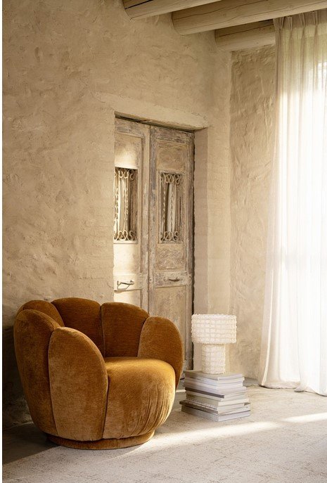 POLTRONA TULIP CATHA6Y - JOLLYGIUNCO Cozy brown rounded chair near a rustic wooden door, with a stacked books and white lamp on the floor in a sunlit room.