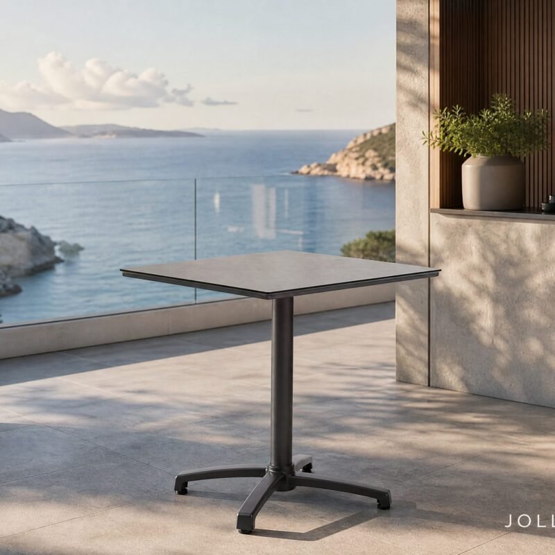 Minimalist outdoor terrace with a grey adjustable-height table, potted plant, and glass railing overlooking the sea. The rocky coastline and distant mountains are in view.