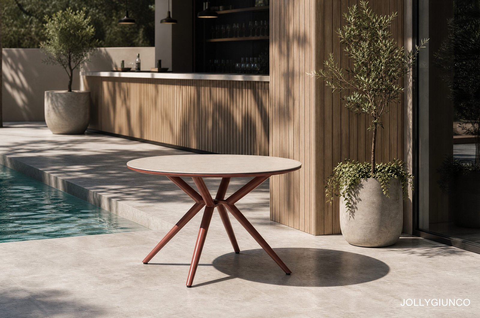 TAVOLO CABAS D120 RAME10 - JOLLYGIUNCO Modern poolside patio with a round pink table, wooden slat wall, and potted olive trees; calm outdoor setting with shadows.