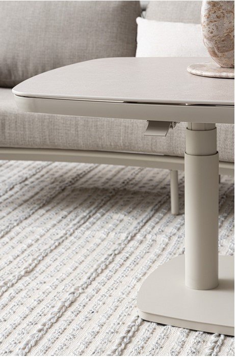 TAVOLO TALETE 135X83X51-72 PELICA5N - JOLLYGIUNCO Close-up of an adjustable-height beige coffee table with a lever under the round tabletop on a light striped rug.